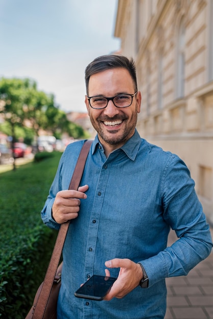 Retrato de profissional sorridente com telefone inteligente em pé na calçada da cidade