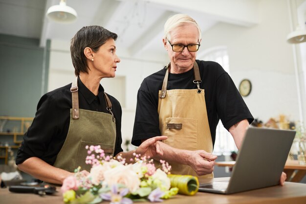Retrato de proprietários de negócios sênior usando laptop juntos enquanto gerenciam o espaço de cópia da floricultura