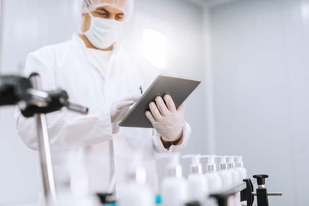 Retrato de químico em uniforme estéril usando tablet em pé ao lado da máquina com sabonetes líquidos