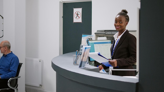 Retrato de recepcionista americano africano trabalhando no balcão de registro no saguão de espera do hospital. Trabalhadora médica usando formulários de relatório de check-up no computador no balcão de recepção.