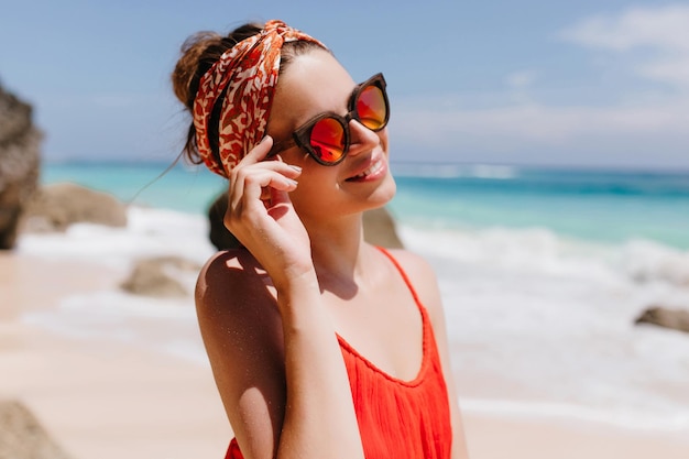 Retrato de rir linda mulher tocando seus óculos de sol de brilho rosa no fundo do mar Adorável modelo feminino bronzeado usa fita no cabelo passando tempo no resort