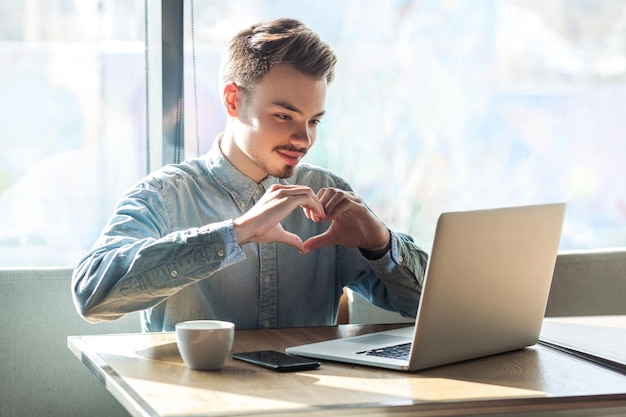 Retrato de romântico jovem namorado feliz na camisa azul está sentado no café e mostrando a sua namorada uma forma de coração com os dedos através de uma webcam online Fundo da janela interna