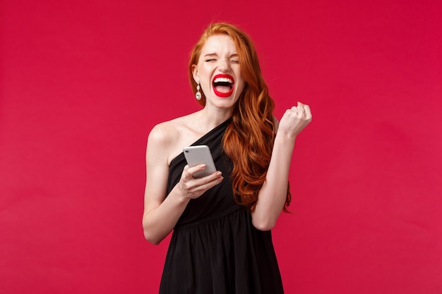 Retrato de ruiva bem-sucedida e satisfeita, ganhando, receba ótimas notícias por mensagem, bomba de presente como campeão, grite sim, ela fez isso, triunfando em vestido preto elegante, parede vermelha