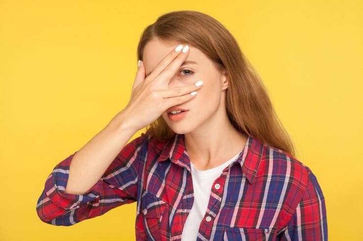 Foto retrato de ruiva curiosa intrometida na camisa quadriculada, cobrindo o rosto e espreitando por entre os dedos com olhar curioso espionando assistindo com interesse tiro de estúdio interior isolado em fundo amarelo