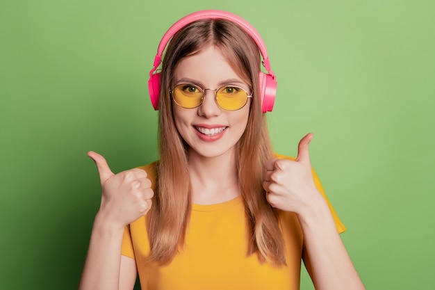 Retrato de senhora fofa mostrar dois polegares para cima aprovar escolha usar fones de ouvido, óculos de sol, camiseta amarela sobre fundo verde