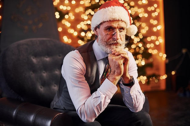Retrato de sênior elegante, com cabelos grisalhos e barba, sentado na sala decorada e com chapéu de Natal.