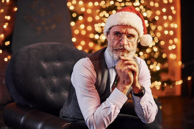 Retrato de sênior elegante, com cabelos grisalhos e barba, sentado na sala decorada e com chapéu de Natal.