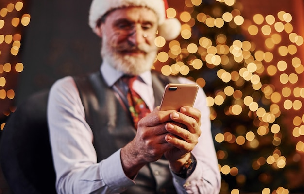 Retrato de sênior elegante, com cabelos grisalhos e barba, sentado na sala decorada e com chapéu de natal.