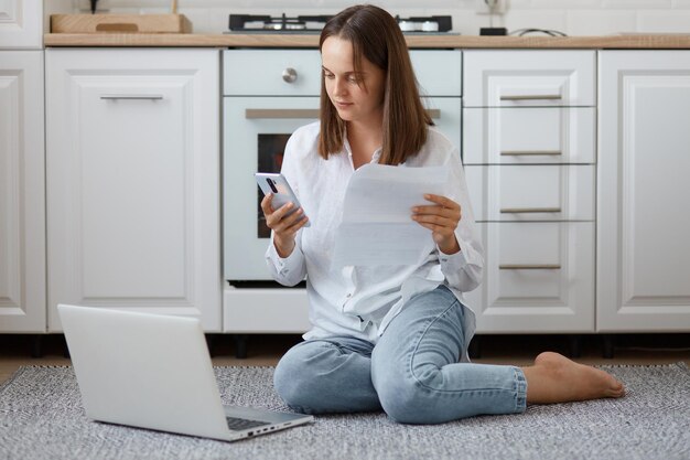 Retrato de sério atraente jovem adulto sentado com o notebook no chão da cozinha e usando o laptop e o telefone inteligente, segurando notas de papel, tentando lidar com os pagamentos online.