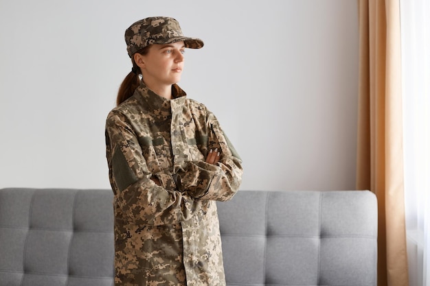 Retrato de sério aviador em pé interno contra o sofá cinza e janela interna, mantendo as mãos postas e olhando para longe, voltando para casa após o saque militar.