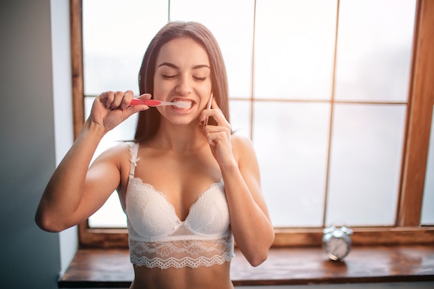 Foto retrato de smiley fresco morena jovem mulher em cueca branca, falando ao telefone enquanto escova os dentes