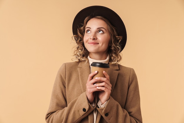 Retrato de sonhar com uma mulher positiva otimista no chapéu isolado na parede bege, segurando a xícara de café.