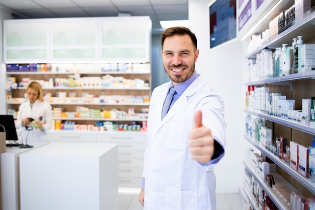 Retrato de sorridente farmacêutico caucasiano em pé na farmácia com os polegares para cima.