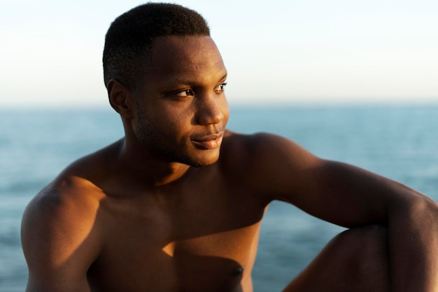 Retrato de sorridente jovem afro-americano sentado na praia e olhando para longe