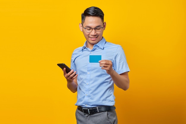 Retrato de sorridente jovem asiático asiático em óculos segurando telefone celular e cartão de crédito isolado no conceito de empresário e empresário de fundo amarelo