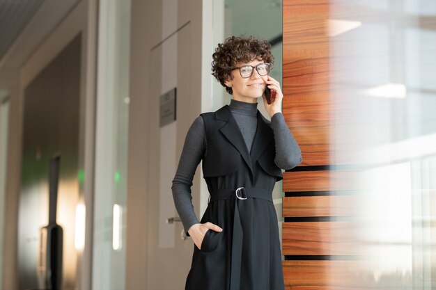 Retrato de sorridente jovem empresária atraente de óculos e vestido preto em pé no corredor e falando com o cliente no telefone