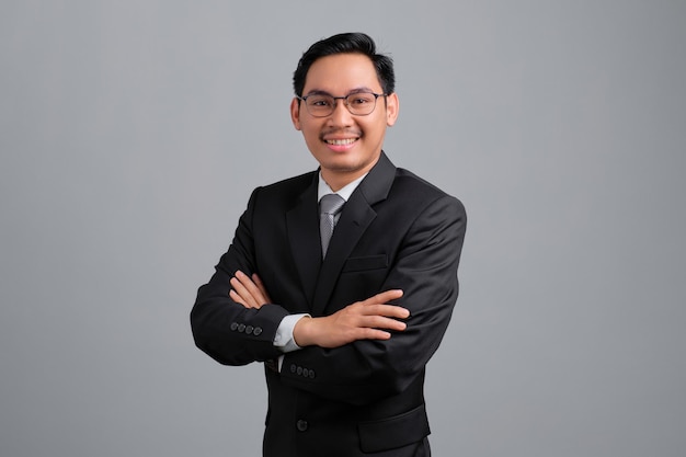 Retrato de sorridente jovem empresário bonito em terno formal e óculos em pé com os braços cruzados e olhando para câmera isolada em fundo cinza