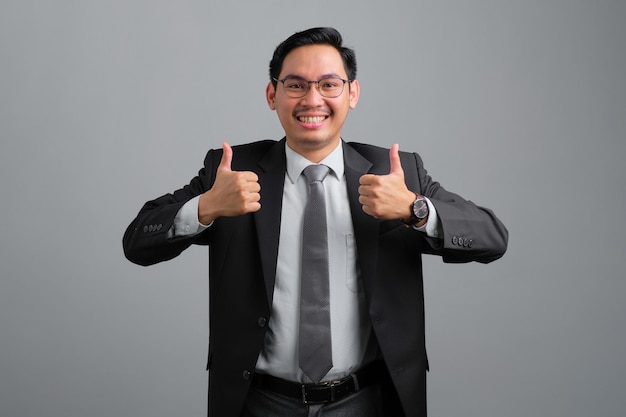 Retrato de sorridente jovem empresário bonito em terno formal, mostrando o polegar para cima isolado no fundo cinza