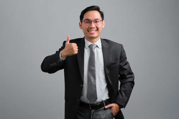 Retrato de sorridente jovem empresário bonito em terno formal, mostrando o polegar para cima isolado no fundo cinza