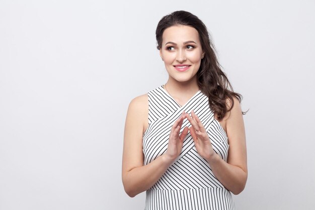 Retrato de sorridente jovem morena bonita com maquiagem e vestido listrado em pé e desviar o olhar com uma cara astuta. tiro de estúdio interno, isolado em fundo cinza.