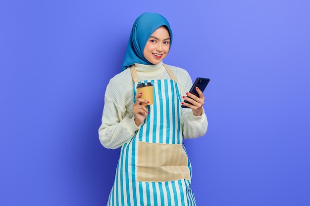 Retrato de sorridente jovem muçulmana asiática em 20 anos vestindo hijab e avental, segurando o telefone celular e o copo de café de papel isolado sobre fundo roxo. Conceito de estilo de vida muçulmano de dona de casa de pessoas