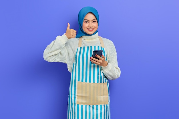 Retrato de sorridente jovem muçulmana asiática vestindo hijab e avental usando smartphone, gesticulando polegares para cima isolado no fundo roxo. Conceito de estilo de vida muçulmano de dona de casa de pessoas