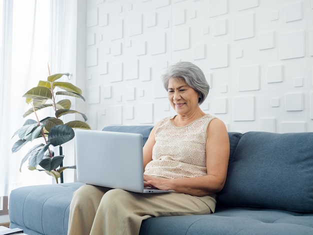 Retrato de sorridente mulher casual sênior asiática sentada no sofá trabalhando com laptop na sala branca brilhante Mulher idosa navega na internet ou videochamada em casa Pessoas mais velhas com tecnologia