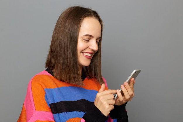 Retrato de sorridente mulher caucasiana alegre com cabelo castanho vestindo suéter usando seu telefone celular conversando com amigos ou lendo postagens em redes sociais em pé isolado sobre fundo cinza