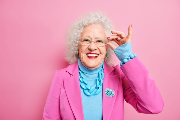 Retrato de sorridente senhora sênior bonita mantém a mão na borda dos óculos usa roupas da moda, feliz em ouvir que algo agradável tem expressão alegre. Moda e estilo de pessoas idosas.