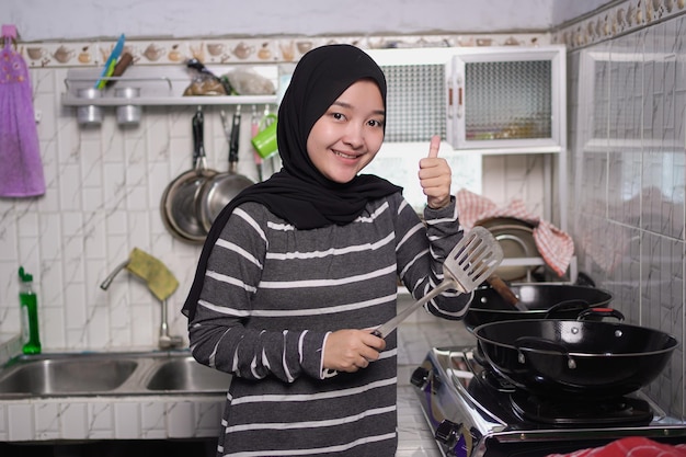 Foto retrato de sorriso atraente mulher muçulmana asiática cozinhando na cozinha vestindo roupas pretas e brancas e segurando uma panela e uma espátula de frente para a câmera polegar para cima gesto vibrações positivas