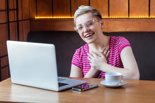 Retrato de sorriso feliz jovem adulta com cabelo curto em camiseta rosa e óculos está sentado com o laptop no café e rindo olhando para a câmera com estilo de vida interior de sorriso