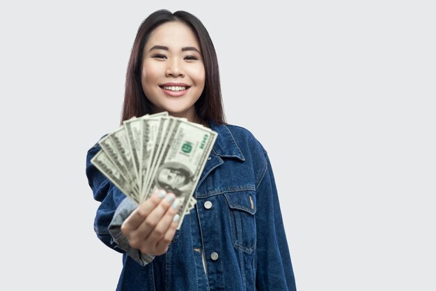 Retrato de sucesso atraente jovem asiática em pé de jaqueta jeans azul, segurando muitos dólares, demonstração e olhando para a câmera com um sorriso. Interior, foto de estúdio, isolado, fundo cinza