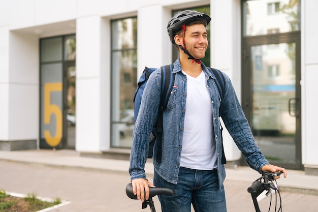 Retrato de tiro médio de alegre bonito jovem entregador em capacete protetor em pé perto de bicicleta na rua da cidade, olhando para longe. mensageiro masculino com mochila térmica entregando comida ao cliente