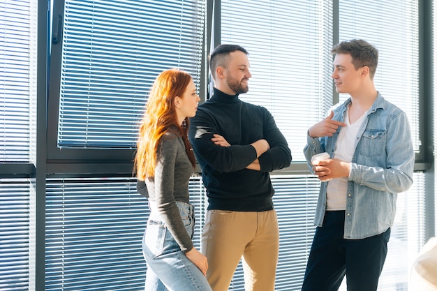 Retrato de tiro médio de três empresários jovens felizes, tendo uma conversa divertida durante a pausa para o café no escritório moderno. Colegas de trabalho confiantes desfrutam de uma comunicação agradável, discutem o projeto pela janela.