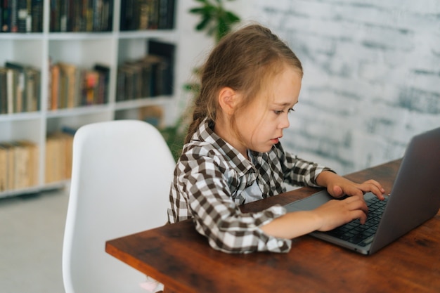 Retrato de tiro médio de uma menina de criança elementar séria usando a digitação no teclado do laptop sentado em