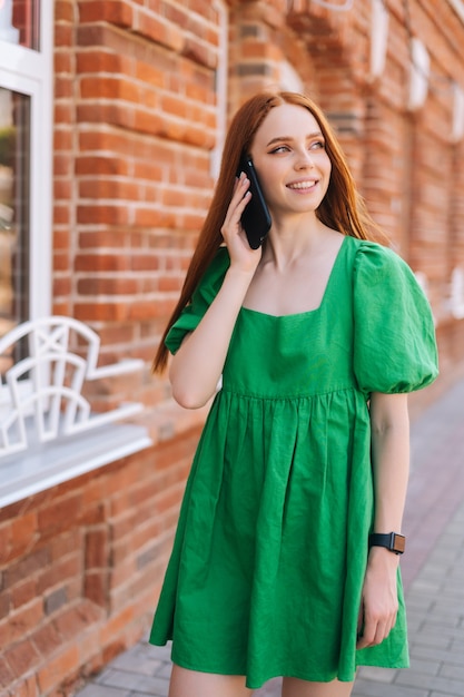 Retrato de tiro médio vertical de mulher jovem feliz falando no celular, andando na rua da cidade em dia de verão. Mulher bonita sorridente usando smartphone ao ar livre, fundo desfocado, foco seletivo