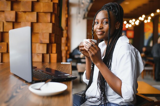 Retrato de tiro na cabeça de uma mulher afro-americana sorridente feliz sentada à mesa no café olhando para a câmera excitada posando trabalhando no computador fazendo lição de casa preparando relatório no café