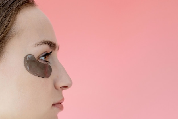 Retrato de tiro na cabeça perto sorridente jovem com máscara de adesivos hidratantes sob os olhos, olhando para a câmera, apreciando o procedimento de cuidados com a pele.