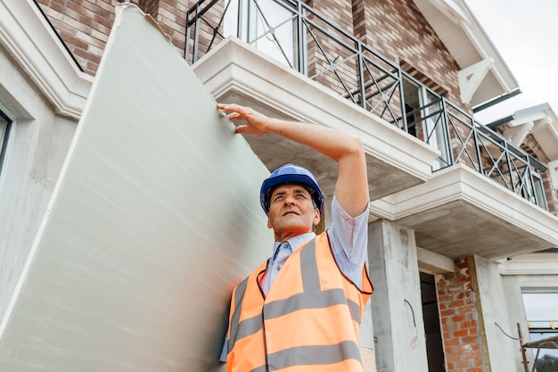 Retrato de trabalhador indiano confiante da construção civil no canteiro de obras segurando nas mãos gerente de construção confiante de drywall de papelão usando capacete de segurança e oeste