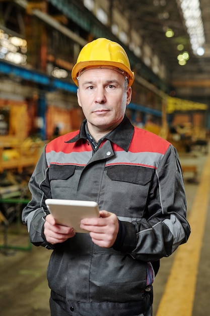 Retrato de trabalhador manual maduro no capacete, olhando para a câmera enquanto trabalhava online no tablet pc na fábrica