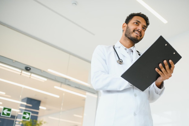 Retrato de trabalhador médico latino indiano amigável feliz vestindo jaleco branco com estetoscópio em volta do pescoço em pé na clínica privada moderna olhando para a câmera Conceito de saúde médica