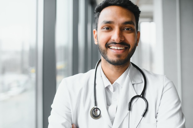 Foto retrato de trabalhador médico latino indiano amigável feliz vestindo jaleco branco com estetoscópio em volta do pescoço em pé na clínica privada moderna olhando para a câmera conceito de saúde médica
