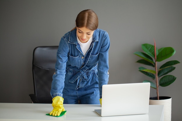 Retrato de trabalhadora feliz limpando a mesa do computador com spray e esponja no escritório