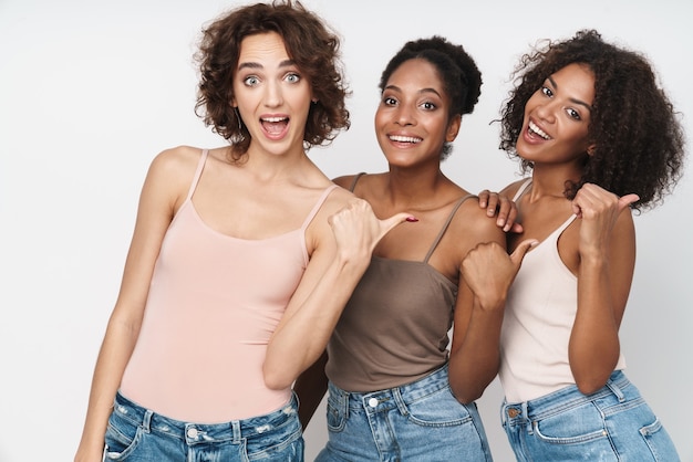 Retrato de três alegres mulheres multiétnicas sorrindo e gesticulando com os dedos isolado na parede branca