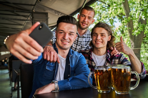 Foto retrato de três amigos do sexo masculino, bebendo cerveja e fazendo selfie no pub.