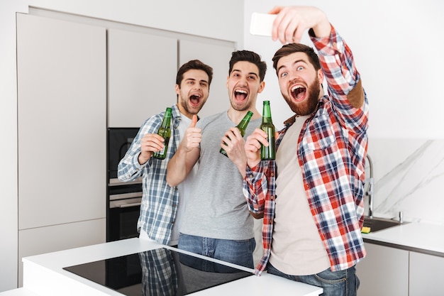 Retrato de três amigos felizes do sexo masculino, comemorando em pé com garrafas de cerveja e tirando uma selfie dentro de casa