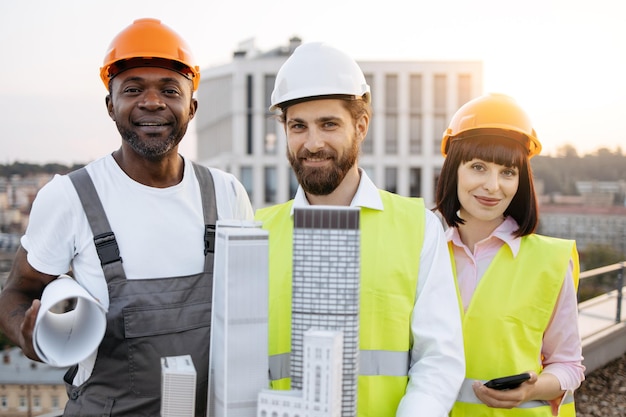 Foto retrato de três designers multiculturais segurando modelos de projetos rolados de edifícios residenciais e smartphones modernos enquanto posam no telhado fundo de paisagem urbana e arranha-céu