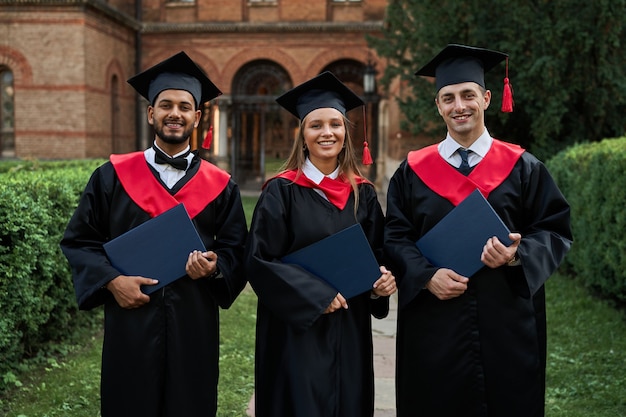 Retrato de três estudantes internacionais femininos e masculinos com diplomas, celebrando a formatura no campus da universidade.