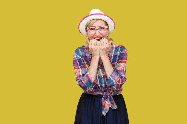 Retrato de triste nervosa moderna elegante mulher madura no estilo casual com chapéu e óculos em pé, roendo as unhas e olhando para a câmera e pânico. estúdio interno tiro isolado em fundo amarelo.