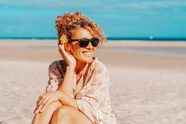 Retrato de turista feliz sentado e sorrindo na praia com céu azul e oceano ao fundo Viagens e turismo nas férias de verão Pessoas do sexo feminino com óculos de sol na areia